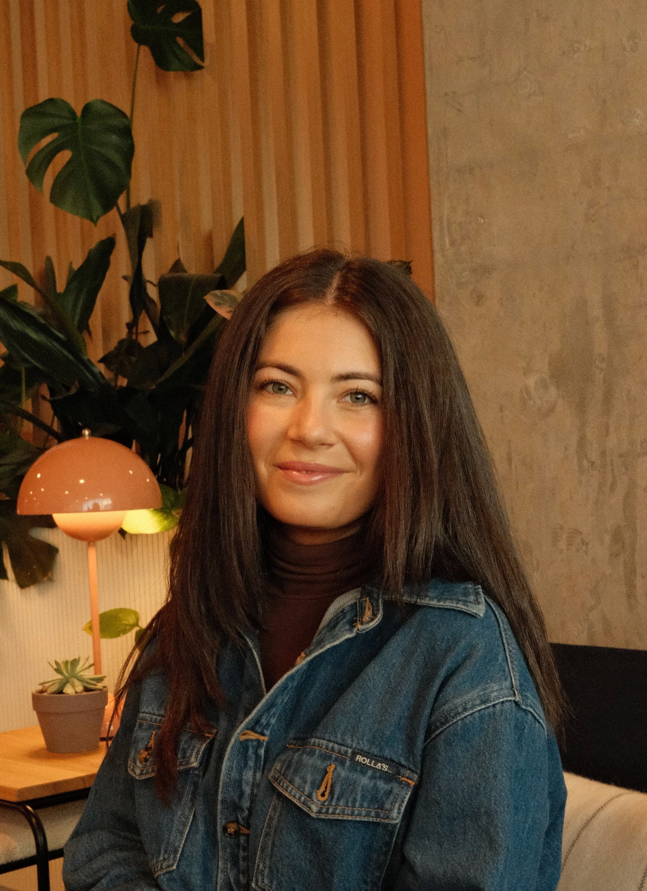 Lauren Cohen, seated in a denim jacket, smiling in a room with a slatted wood wall and leafy plants.